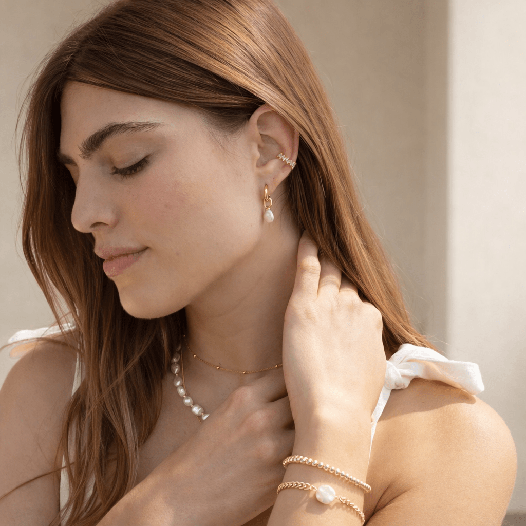 Woman wearing gold jewelry including earrings, necklace, and bracelets against a neutral background